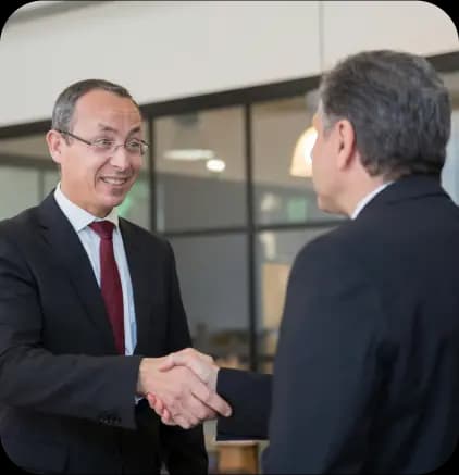 Two businessmen in suits shaking hands. One man on the left is smiling and wearing glasses, while the other man's back is to the camera.
